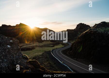 Curvy asphalt road going through mountainous terrain against sunset sky with bright sun in evening in nature