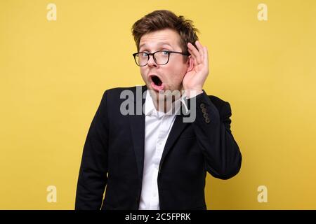 Handsome caucasian guy trying to secretly listen in on a conversation, latest gossips isolated on yellow background Stock Photo