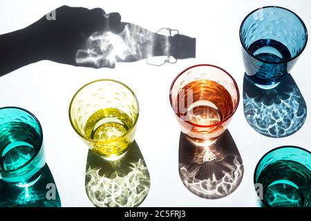 Shadow of a hand holding a bottle that pours water into colored glass cups under the strong sunlight that casts its textured, colored shadows on a whi Stock Photo