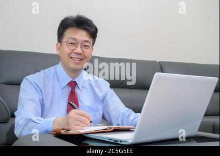 Middle-aged Asian businessman sitting on sofa in living room working at home. Telecommuting concept. Stock Photo