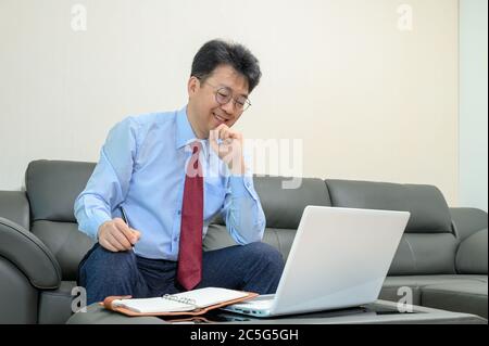 Middle-aged Asian businessman sitting on sofa in living room working at home. Telecommuting concept. Stock Photo