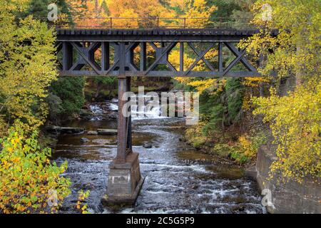 Railroad bridge over a river with a waterfall in the background surrounded by vibrant fall colors in a small Upper Peninsula town of Michigan. Stock Photo