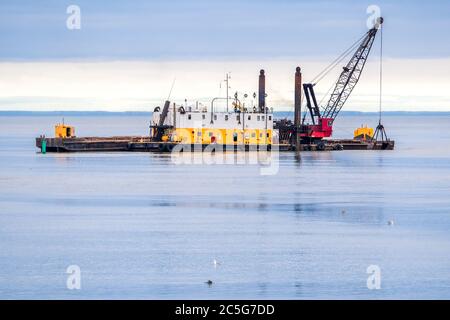 An old dredging barge in the ocean. Barge is yellow and white with a red crane on the deck. Scoop is being lowered into the water. Dark overcast sky. Stock Photo