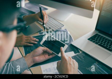 Software developers are using smart phones and analyzing together the code written in the program on the computer. Stock Photo