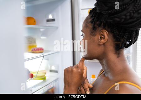 African Woman Think About Food Near Fridge In Kitchen Stock Photo