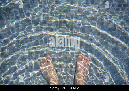 White man's feet in the pool, mixed dark and light gray mosaic floor mixed The waves on the water surface jump in the golden sun. Stock Photo