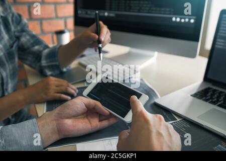 Software developers are using smart phones and analyzing together the code written in the program on the computer. Stock Photo