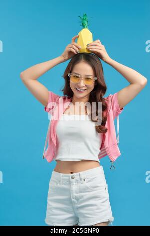 excited Asian girl holding pineapple over her head and smiling at camera isolated on blue background Stock Photo