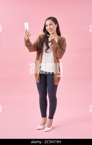 Happy young asian woman casually dressed standing isolated over pink background, using mobile phone Stock Photo