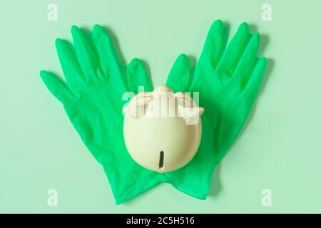 Piggy bank on green latex gloves isolated on light green background flat lay. Top view of piggy bank on rubber gloves. Concept of financing and saving Stock Photo