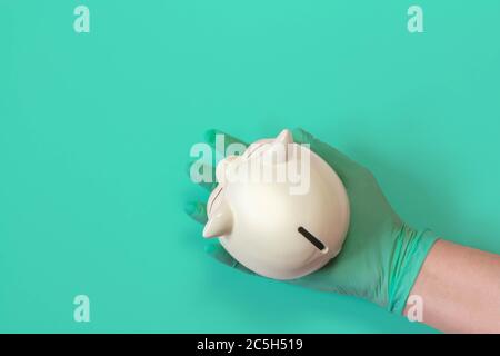 Hand in green latex glove holding piggy bank on green background with copy space. Top view of hand in glove with piggy bank. Concept of financing and Stock Photo