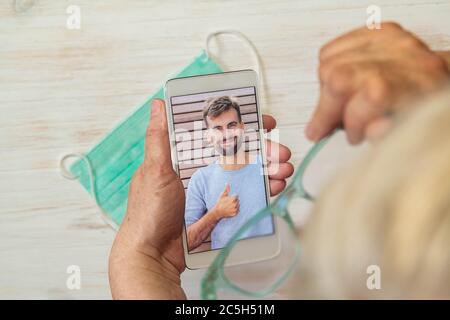 Female with white hair and glasses looking at guy in phone, medical face mask on table. Guy making video call to mom. Senior woman making video call Stock Photo