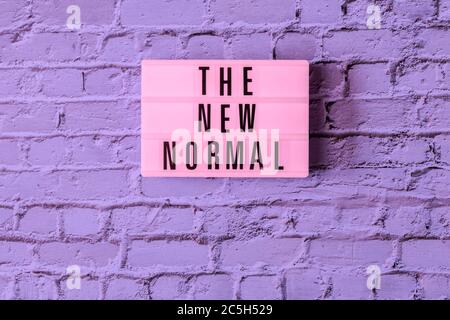 THE NEW NORMAL sign hanging on wall in neon colors. Pink signboard THE NEW NORMAL in on vintage purple brick wall background in fluorescent light. Stock Photo