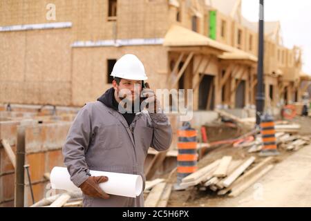 Foreman speaking by smartphone and holding drawing plan at construction site. Stock Photo