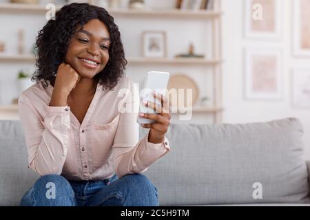 Cool App. Pretty black woman sitting on couch and using smartphone Stock Photo