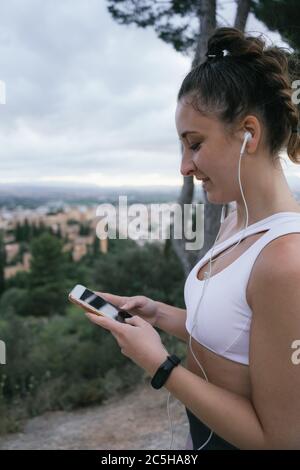 Sporty young woman in headphones with phone. Pretty fit woman using the smartphone after exercising in a viewpoint of the Alhambra on a cloudy summer. Stock Photo