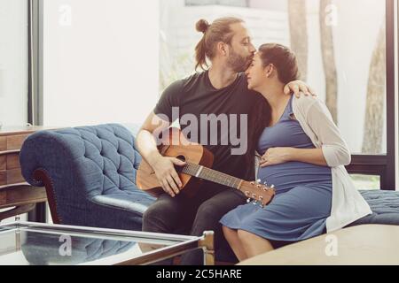 Husband with pregnant wife stay home lovely playing guitar music for baby kissing and showing love together Stock Photo