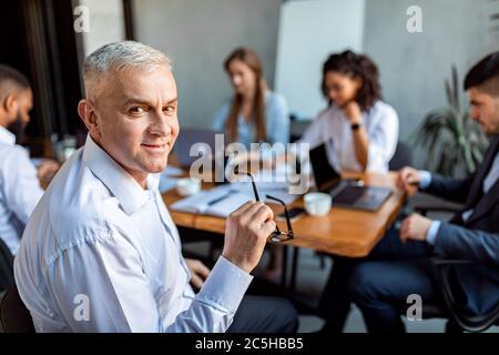 Senior Businessman Sitting At Corporate Meeting With Employees In Office Stock Photo