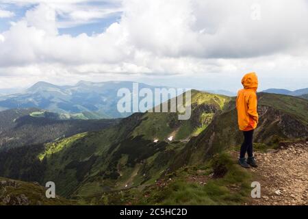 Tourist girl in orrange coat looks on the trail along the mountain range. Hiking travel outdoor concept panoramic view. Stock Photo