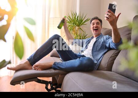 Man smiling drinking a beer from a bottle and having snack sitting on the sofa in the living room at home Stock Photo