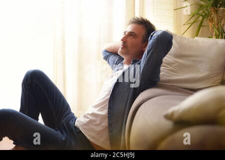 Pensive man sitting on the floor and leaning on a sofa in the living room at home Stock Photo