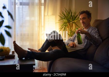 Relaxed man entertaining with a laptop at home having a beer sitting on a sofa Stock Photo