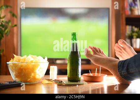 Man at home watching game on television with feet on side table and snack. Stock Photo