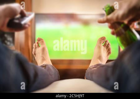 man sitting on a sofa watching match on TV with remote control and beer at home Stock Photo