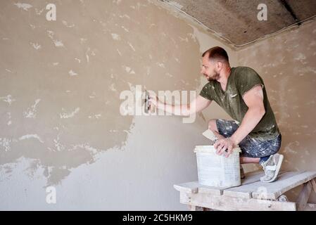 Young man at wooden stand is puttying the walls indoors. Guy with beard in t-shirt and jeans is smeared with putty. Small room with gray walls Stock Photo