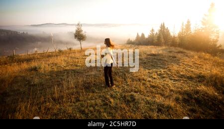Carefree happy woman in yellow shirt and straw hat enjoying nature on grass meadow on top of mountain with sunrise. Beauty girl outdoor with sunbeams. Stock Photo