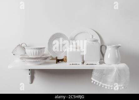 Composition of white wood hanging shelf on wall with country home tableware set on display. Romantic pattern plates, ceramic storage box containers. Stock Photo