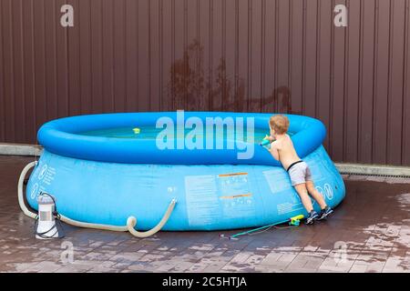 Cute little adorable caucasian blond toddler boy looking into inflatable blue pool enjoy playing with toy fishing rod at home yard on hot summer day
