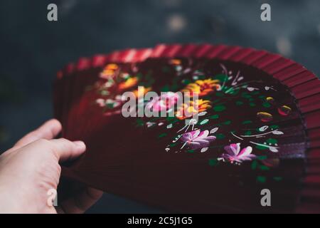 Hand holding red wooden folding hand fan with floral pattern on dark background. Selective focus. Copy space. Stock Photo