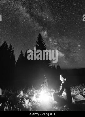 Side view of female tourist having a rest by campfire on background of spruce forest under starry sky on which milky way is visible. Bonfire with a big flame under the night sky. Monochrome image Stock Photo