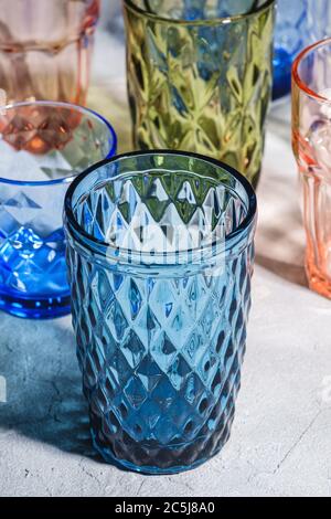 Colorful faceted and geometric drink glass, group of empty green, red, blue and transparent drinkware for beverage on stone concrete background, angle Stock Photo