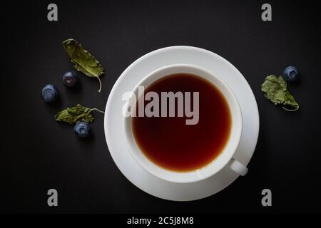 White cup with tea, blueberries and dried mint leaves on a dark background, view from above Stock Photo