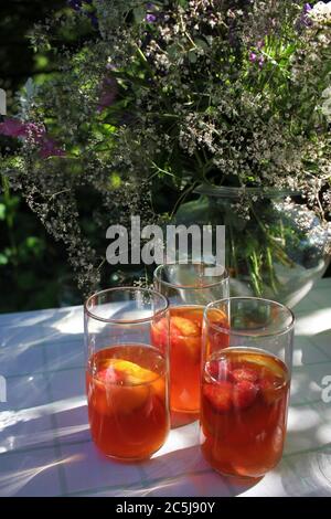 Glass ow white sangria and summer flowers on the cloth in the garden. White wine with fruits, oranges, strawberries and peaches in the glasses Stock Photo