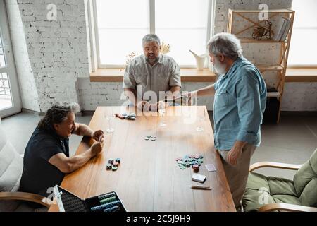 Always young. Group of happy mature friends playing cards and drinking wine. Look delighted, excited. Caucasian men gambling at home. Sincere emotions, wellbeing, facial expression concept. Stock Photo