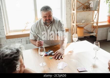 Young at heart. Two happy mature friends playing cards and drinking wine. Look delighted, excited. Caucasian men gambling at home. Sincere emotions, wellbeing, facial expression concept. Smiling. Stock Photo