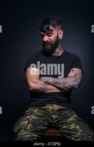 Brutal attractive bearded man with tattooed hands poses in black blank t-shirt and army pants seated on a chair Stock Photo