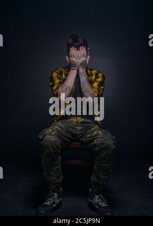 Picture on a black background of a bearded male model sitting on a chair and covering his face with his tattooed hands Stock Photo