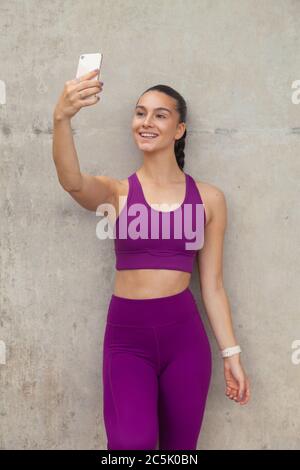 An attractive woman wearing sportswear leaning against a concrete wall and holding up a mobile phone. Stock Photo