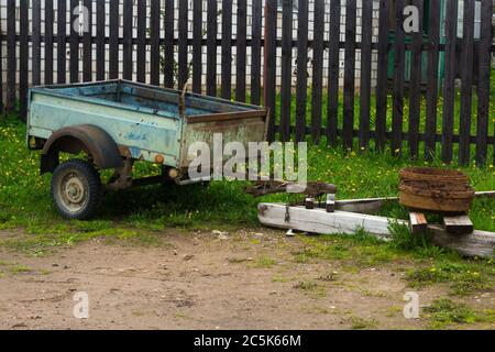 an old blue trailer stands in a village, old rusty rims hold it. against the background of the fence Stock Photo