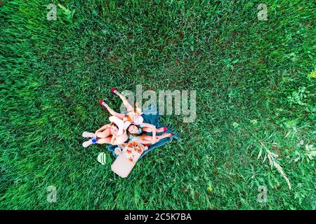 Mother and two daughters have a picnic with pizza on a green grass, aerial top view Stock Photo