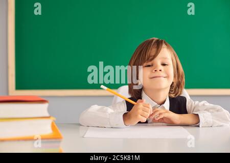 Back to school. Cute little boy schoolboy smiling while sitting at a table on the background of the school blackboard. Stock Photo