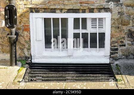 Newly painted basement window seen from the adjacent pavement. A metal grating is seen, used for drainage during heavy rain. Stock Photo