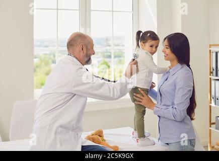 Family with a visit to the doctor in the clinic office. Senior doctor with a stethoscope listens to a little girl with mother patients in the hospital Stock Photo