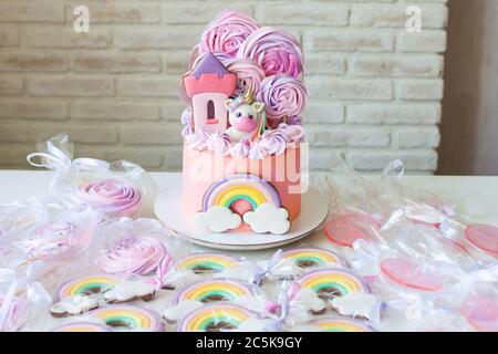 Pink birthday cake with fondant unicorn, rainbow and meringue clouds. Candy bar with cake, cookies and lollipops. Stock Photo