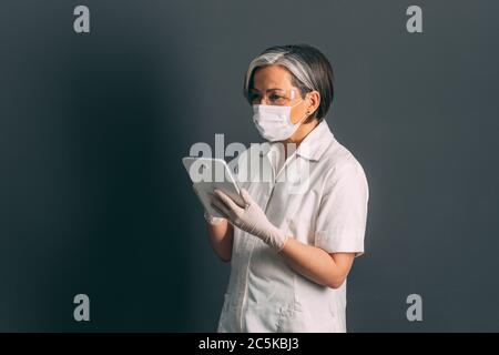 Experienced female doctor using tablet computer. Mature Caucasian woman in protective uniform working with digital gadget. Advanced technology in Stock Photo