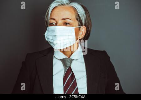 Gray-haired business woman wearing protective mask and office formalwear looking at side. Close up portrait on gray back. Tinted image Stock Photo